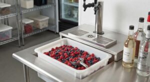A commercial bar prep station featuring a white plastic bin filled with fresh raspberries and blueberries, accompanied by a metal scoop, a tap draft tower, and bottles of simple syrup and vodka.
